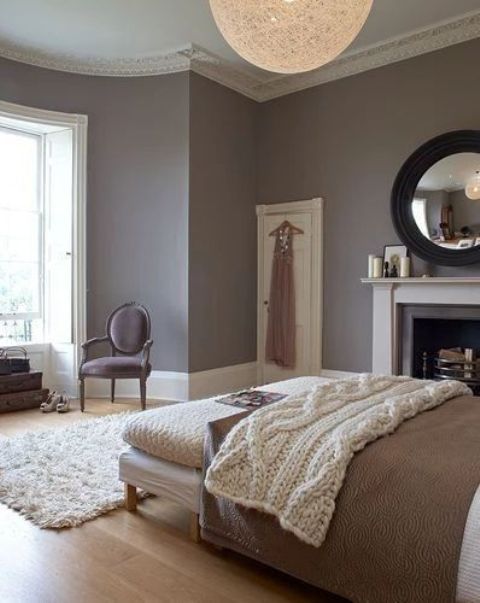 a refined taupe bedroom with molding, a neutral bed and bench, cozy blankets, a chic chair and a vintage fireplace