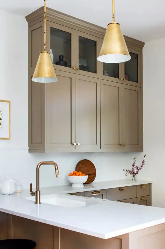 a refined taupe kitchen with shaker cabinets - mostly lower and a couple of upper ones, white stone countertops and a backsplash, gold pendant lamps