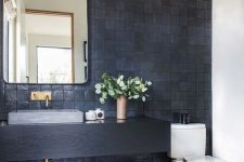 a stylish bathroom with a black Zellige tile wall, a black vanity, a rectangular mirror, a pendant lamp and a grey stone sink