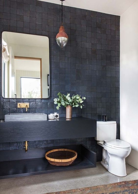 a stylish bathroom with a black Zellige tile wall, a black vanity, a rectangular mirror, a pendant lamp and a grey stone sink