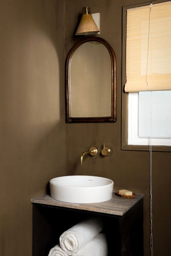 a taupe powder room with a black floating vanity, a round white sink, an arched mirror and a woven shade