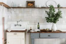 a vintage kitchen with white Zellige tiles and a planked floor, a neutral vanity and a grey kitchen island, a long stained open shelf instead of cabinets