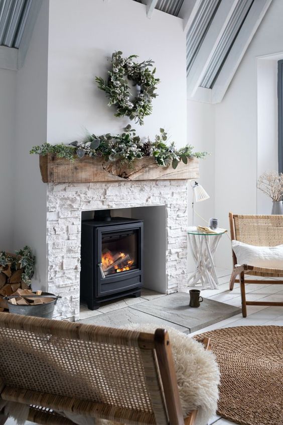 a welcoming neutral space with a wood burning stove, a rough wood slab mantel, greenery for winter decor, cane furniture and firewood in buckets