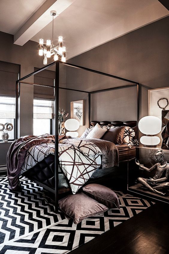 a whimsical bedroom with taupe walls and Roman shades, a black canopy bed with printed bedding, geometric rugs and catchy lamps