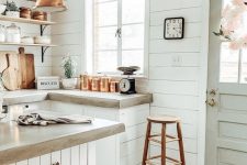 a white famrhouse kitchen clad with shiplap and shiplap cabinets, concrete countertops, a copper pendant lamp and a tall stool