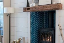 a white planked wall, a fireplace clad with navy herringbone tiles and a hearth placed inside it and a wood slab mantel