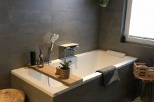 an elegant bathroom clad with taupe tiles, a bathtub clad with them, some potted greenery, baskets and crates for storage
