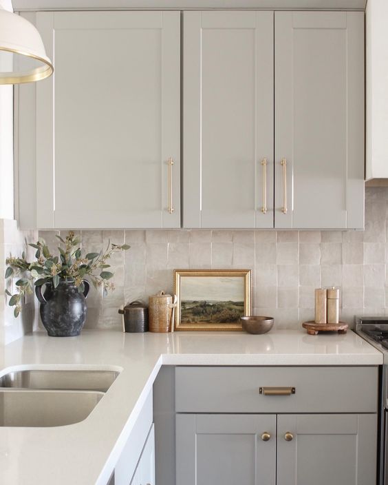 an elegant dove grey kitchen with shaker style cabinets, white stone countertops, a neutral Zellige tile backsplash and brass fixtures