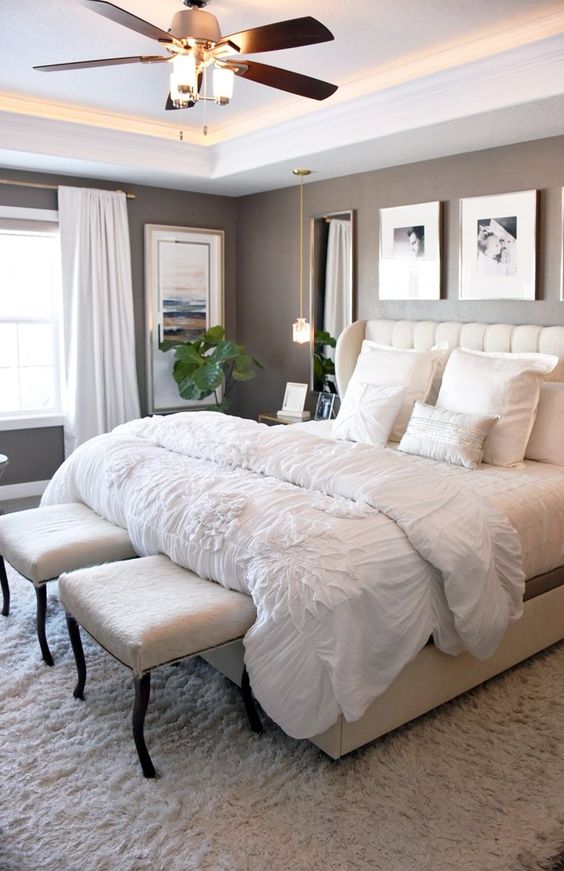an elegant master bedroom with taupe walls, a large creamy bed, neutral stools, a stained dresser, mirrors and a lit up ceiling