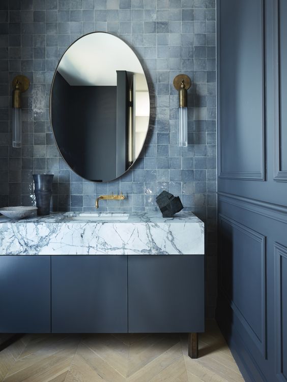 an elegant stone blue bathroom with a matching vanity, a blue Zellige tile backsplash, an oval mirror and cool brass sconces