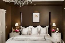 an elegant taupe bedroom with a black bed with pillars, white bedding, white nightstands, a chic crystal chandelier