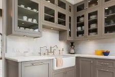an elegant taupe vintage kitchen with shaker style cabinets, glass upper ones, a white stone countertop and a backsplash and vintage fixtures and knobs is all cool