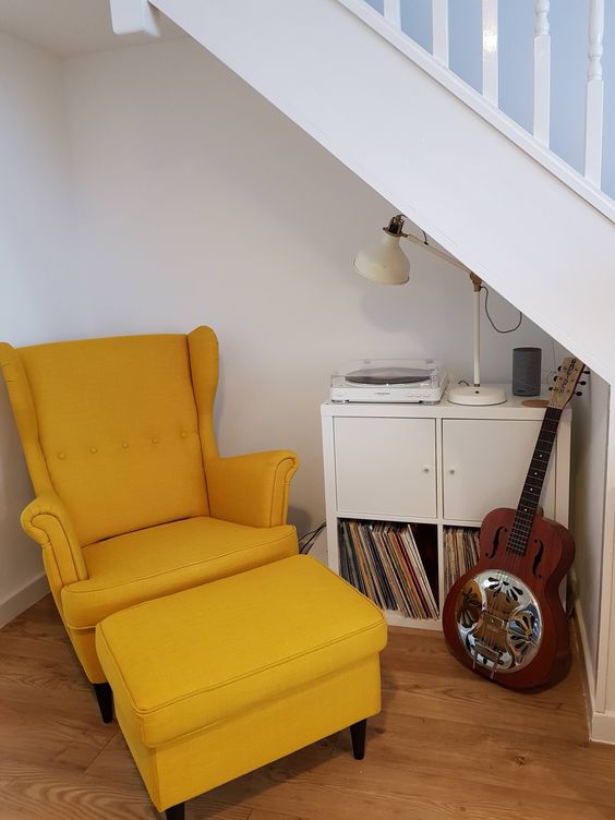 a small and stylish reading corner under the stairs, with a chic vinyl cabinet, a guitar and a mustard chair with a footrest