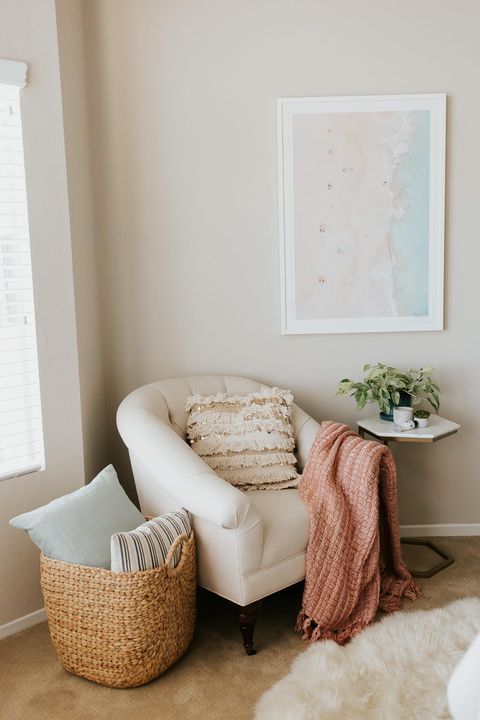 a small and lovely reading nook with a creamy chair, a couple of pillows and a blanket, a beach artwork, a small table and a faux fur rug