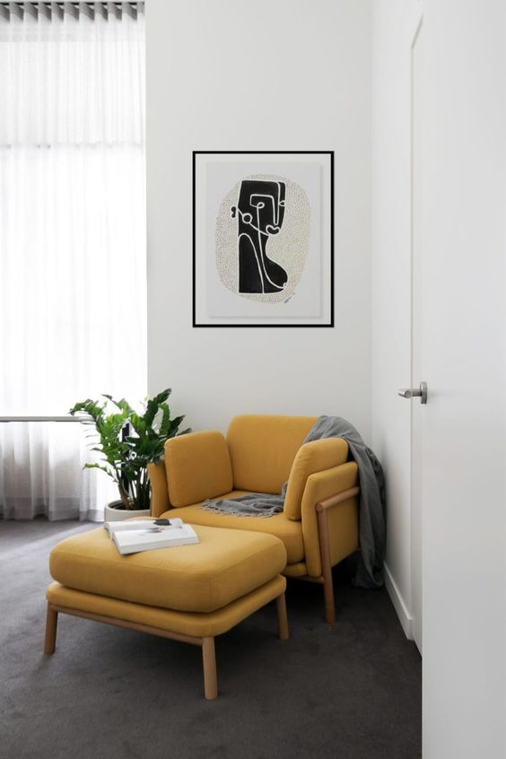 a modern sitting corner with a pretty artwork, a mellow yellow chair and a footrest, a blanket and a potted plant