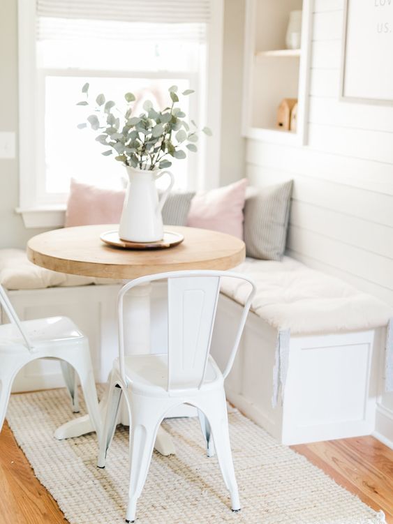a white farmhouse breakfast corner with a built in bench with storage, a round table, white metal chairs and built in niches for storage