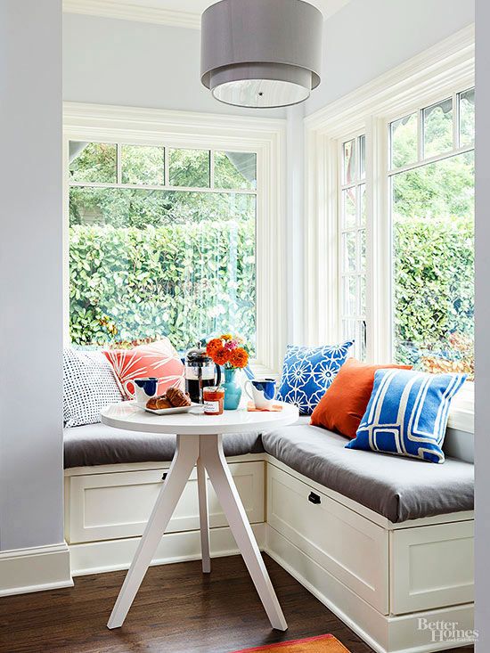 a super cozy window nook with a built-in storage bench with pillows, a round table and a grey pendant lamp plus lots of light