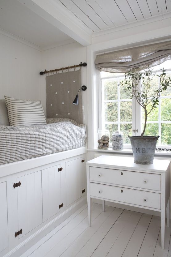 a lovely white cottage bedroom with a bed placed on a large storage unit, a dresser as a nightstand and grey textiles