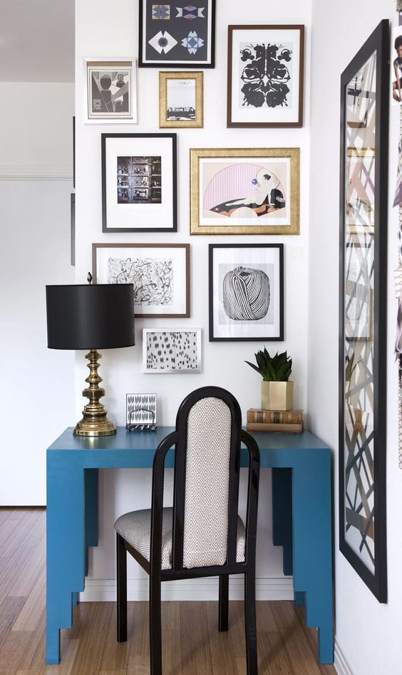 a small space with a beautiful gallery wall. a bold blue desk, an elegant art deco chair and a chic black and gold lamp