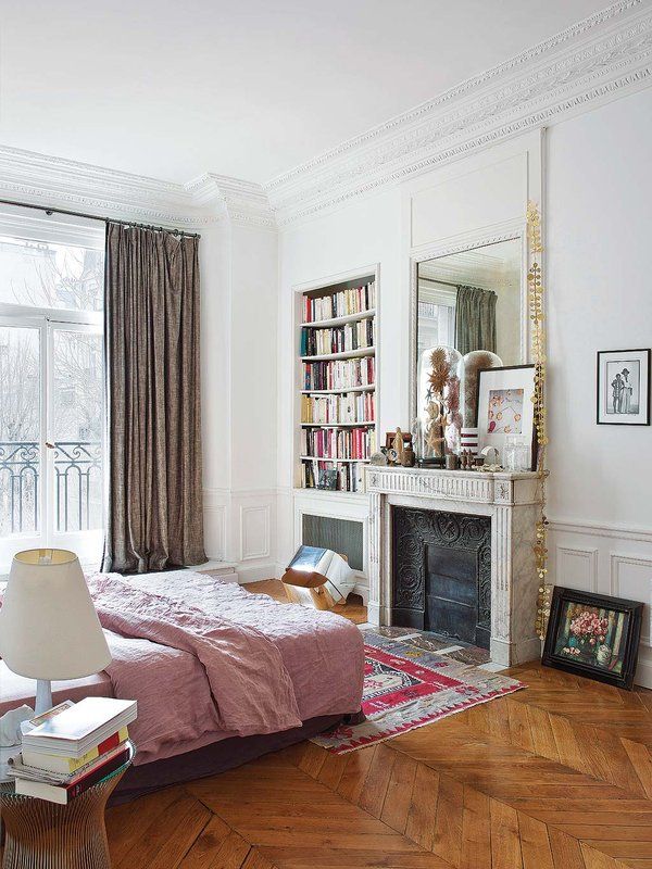 a French chic bedroom with paneling, an antique non-working fireplace, a bed with pink bedding, built-in shelves and a mirror