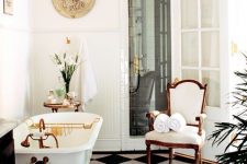 a beautiful Parisian style bathroom with black and white tiles on the floor, a white clawfoot tub, a vintage chair, a chic chandelier and some blooms