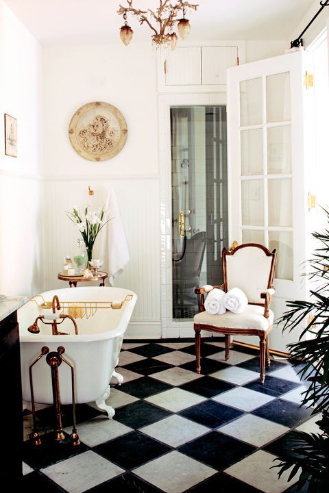 a beautiful Parisian style bathroom with black and white tiles on the floor, a white clawfoot tub, a vintage chair, a chic chandelier and some blooms