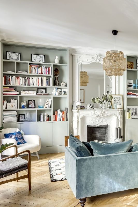 a beautiful and light filled French chic living room with grey bookcases, a vintage fireplace, a slate blue sofa and some pretty modern chairs