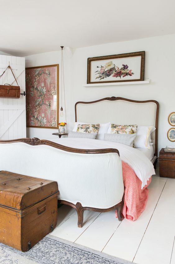 a beautiful cottage bedroom in creamy shades, with a vintage bed with neutral bedding, bold art, a mini table and a stained chest as nightstands