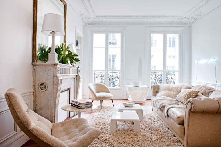 a beautiful neutral living room with an antique fireplace, a tan sofa and tan chairs, white stools and some elegant lamps