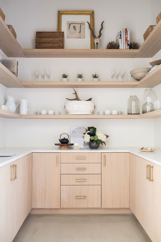 a blonde wood kitchen with sleek cabinets and long open shelves over them, white stone countertops and potted greenery and blooms