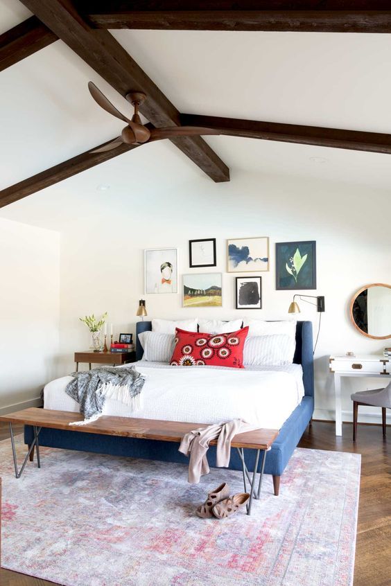 a bold and chic bedroom with dark stained beams, a navy upholstered bed, a woooden bench, a bright gallery wall , a white vanity and a stained table as nightstands