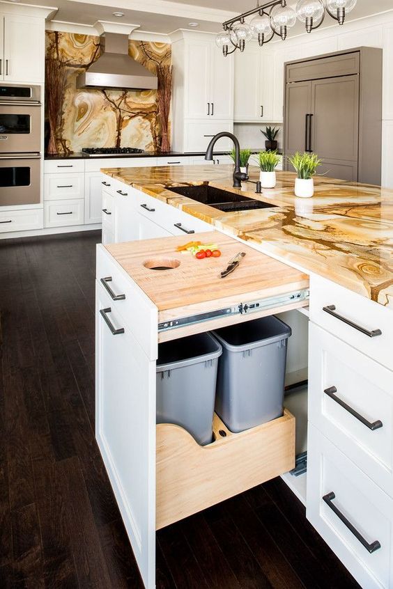 a bold kitchen with white farmhouse cabinets, tan and brown stone countertops and a backsplash, a kitchen island with lots of drawers