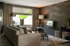 a catchy and stylish living room with an accent taupe wall, tan curtains and a sofa, a tan rug, tan leather poufs and a black TV unit
