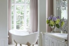a classic French bathroom in neutrals, with a clawfoot tub by the window, lilac curtains and a large vanity, crystal lamps and a chandelier