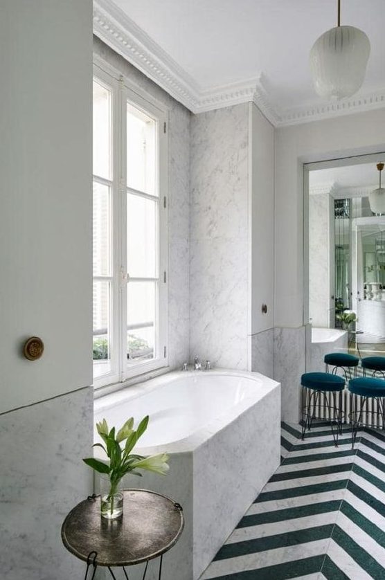 a contemporary French chic bathroom done with white marble, chevron clad floor, teal stools and cabinets on the walls