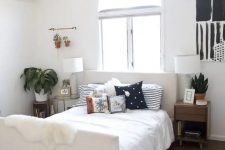 a cool boho bedroom with a creamy bed, black and white pillows, an elegant glass table and a stained nightstand, potted plants and a blakc and white artwork