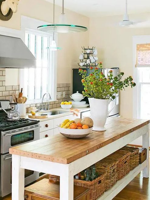 a cozy cottage kitchen with white cabinets, butcherblock countertops, glass pendant lamps, stainless steel appliances and a table kitchen island with an open shelf and blooms
