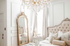 a creamy French chic bedroom with paneling, a blush tufted bed with a vintage design, a floor mirror, a fantastic crystal chandelier