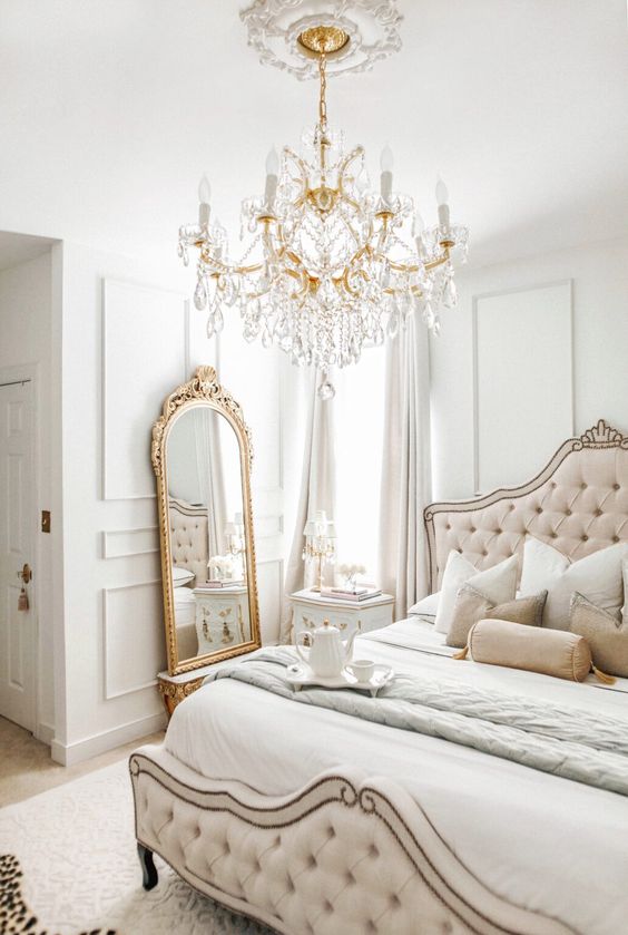 a creamy French chic bedroom with paneling, a blush tufted bed with a vintage design, a floor mirror, a fantastic crystal chandelier