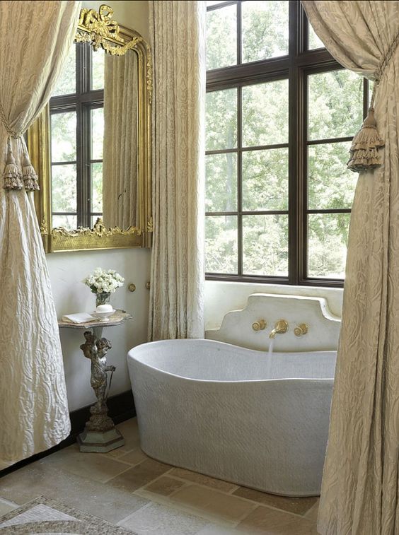 a fabulous French bathroom with a large window, a neutral bathtub, a large mirror in a gold frame, neutral curtains and a tiled floor