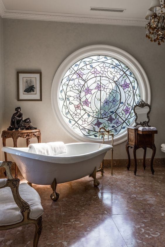 a fantastic French chic bathroom with grey walls, a mosaic round window, a clawfoot tub, a vintage vanity and a side table plus a vintage chair