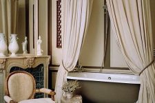 a fantastic neutral vintage-inspired Parisian bathroom with canopies, a bathtub, a vintage chair and a faux fireplace