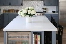 a grey farmhouse kitchen with white glossy herringbone tiles, white countertops, an oversized grey and white kitchen island with  open storage shelves and a drink cooler
