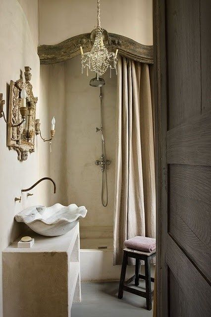 a jaw dropping vintage French bathroom with tan walls, a vanity of stone and a shower space, a mirror in an ornated frame and a crystal chandelier