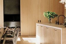 a large and cozy blonde wood kitchen with sleek cabinets, a herringbone floor, leather woven chairs and greenery