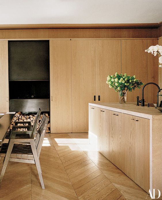 a large and cozy blonde wood kitchen with sleek cabinets, a herringbone floor, leather woven chairs and greenery
