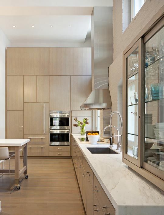 a large kitchen with blonde wood cabinetry, white marble countertops, windows instead of a backsplash and stainless steel appliances