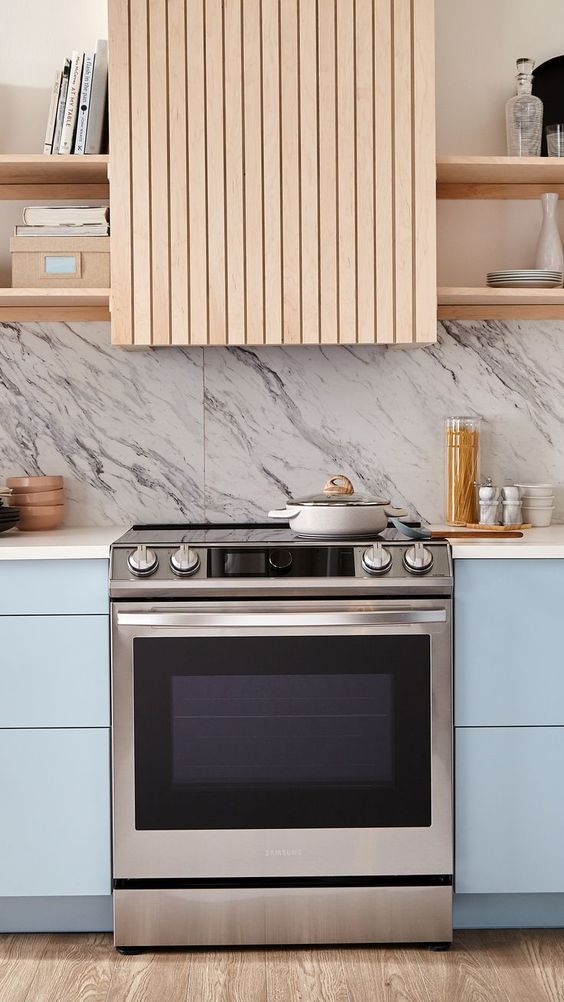 a light blue kitchen with sleek cabinetry, a white marble backsplash, open blonde wood shelves and a hood clad with matching wooden slabs