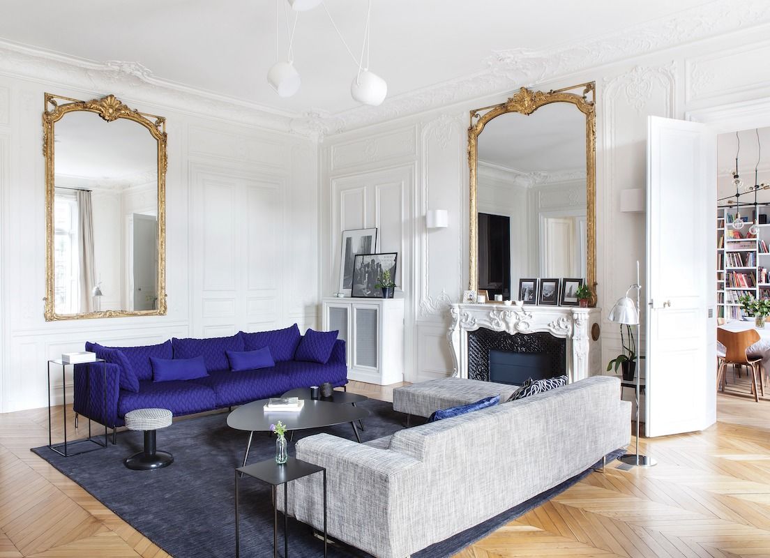 a lovely modern Parisian living room with an antique fireplace, a bold purple and grey sofa, a bench and some table and a couple of mirrors