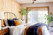 a lovely rustic bedroom with wooden walls and a ceiling, a metal bed with bright bedding, modenr dressers as nightstands and a vintage lamp